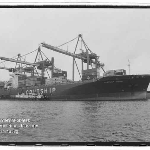 Schwarzweißaufnahme eines großen Containerschiffs mit der Aufschrift CONTSHIP, das am Hafen von mehreren Containerkränen be- oder entladen wird, vor ruhigem Wasser.
