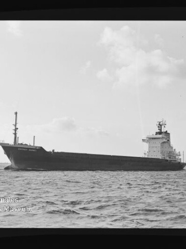 Großes Frachtschiff seitlich auf offener See mit achtern liegender Brücke und Mast am Bug, ruhige, leicht wellige See, bewölkter Himmel und ein weiteres Schiff am Horizont; Wasserzeichen des Internationalen Maritimen Museums Hamburg unten links.