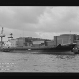 Schwarzweißaufnahme eines am Kai liegenden Frachtschiffs mit der Aufschrift „CMC“, im Hintergrund Lagerhallen und ein Kran, im Vordergrund Wasser sowie sichtbarer Filmrand und Wasserzeichen des Internationalen Maritimen Museums Hamburg.