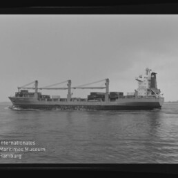 Schwarzweiß-Seitenaufnahme eines Frachtschiffs mit gestapelten Containern und drei Ladekränen, das auf ruhigem Wasser fährt; am Bildrand sichtbarer Filmrahmen und Museum-Logo (Internationales Maritimes Museum Hamburg).