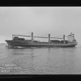Schwarzweißaufnahme eines seitlich sichtbaren Frachtschiffs mit drei Ladekränen und auf Deck gestapelter Ladung, das auf ruhigem Wasser fährt; Filmrand und Wasserzeichen des Internationalen Maritimen Museums Hamburg sind sichtbar.