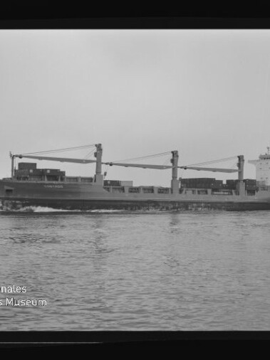 Schwarzweißaufnahme eines seitlich sichtbaren Frachtschiffs mit drei Ladekränen und auf Deck gestapelter Ladung, das auf ruhigem Wasser fährt; Filmrand und Wasserzeichen des Internationalen Maritimen Museums Hamburg sind sichtbar.