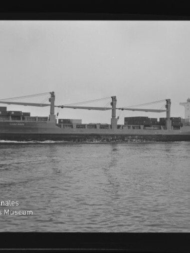 Schwarzweiß-Foto einer Seitenansicht eines Frachtschiffs mit drei großen Ladekränen und auf dem Deck gestapelten Containern, das ruhig über dem Wasser fährt; am unteren linken Rand das Logo des Internationalen Maritimen Museums Hamburg und Rand des Negativfilms sichtbar.