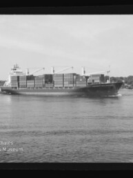 Schwarzweißaufnahme eines Containerschiffs mit gestapelten Ladecontainern und Deckkränen, das auf einem ruhigen Fluss vor einem bewaldeten Ufer fährt; unteren linken Bildrand ziert das Logo des Internationalen Maritimen Museums Hamburg.