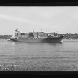 Schwarzweißaufnahme eines Containerschiffs mit gestapelten Ladecontainern und Deckkränen, das auf einem ruhigen Fluss vor einem bewaldeten Ufer fährt; unteren linken Bildrand ziert das Logo des Internationalen Maritimen Museums Hamburg.