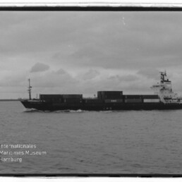Schwarzweißaufnahme eines Containerschiffs in Seitenansicht auf ruhiger See unter bewölktem Himmel, mit gestapelten Containern und Aufbauten am Heck; Logo des Internationalen Maritimen Museums Hamburg unten links sichtbar.