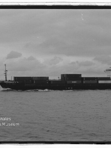 Schwarzweißaufnahme eines Containerschiffs in Seitenansicht auf ruhiger See unter bewölktem Himmel, mit gestapelten Containern und Aufbauten am Heck; Logo des Internationalen Maritimen Museums Hamburg unten links sichtbar.