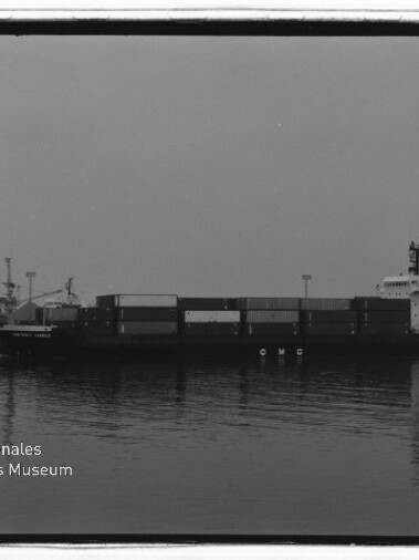 Schwarzweißaufnahme eines Containerschiffs mit gestapelten Containern, das ruhig im Hafenwasser liegt und sich spiegelt; unten links das Logo des Internationalen Maritimen Museums Hamburg.