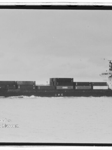 Schwarzweiß-Aufnahme eines Containerschiffs in Seitenansicht mit gestapelten Seecontainern auf ruhiger See; unten links sichtbar das Wasserzeichen des Internationalen Maritimen Museums Hamburg.