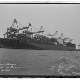 Schwarzweißfoto eines großen Containerschiffs am Kai, beladen mit gestapelten Containern und umgeben von mehreren Hafenkränen, aufgenommen vom Wasser aus; links unten das Signet des Internationalen Maritimen Museums Hamburg.