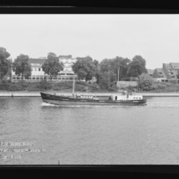 Schwarzweißfoto eines schmalen Frachtschiffs mit kleinem Aufbau und Mast, das auf einem Fluss eine leichte Bugwelle zieht; im Hintergrund Bäume und mehrgeschossige Gebäude am Ufer, links unten sichtbar das Wasserzeichen des Internationalen Maritimen Museums Hamburg und der schwarze Negativrand des Films rahmt das Bild.