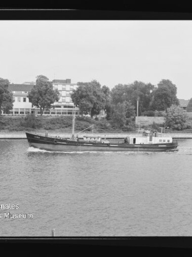Schwarzweißfoto eines schmalen Frachtschiffs mit kleinem Aufbau und Mast, das auf einem Fluss eine leichte Bugwelle zieht; im Hintergrund Bäume und mehrgeschossige Gebäude am Ufer, links unten sichtbar das Wasserzeichen des Internationalen Maritimen Museums Hamburg und der schwarze Negativrand des Films rahmt das Bild.