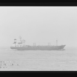 Großes Frachtschiff fährt bei diesigem, nebeligen Wetter auf ruhiger See, mit sichtbarer Bugwelle; Foto zeigt außerdem Filmrandmarkierungen und ein Wasserzeichen des Internationalen Maritimen Museums Hamburg.