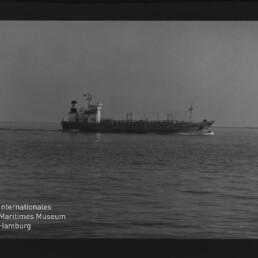 Schwarzweißaufnahme eines mittig platzierten Frachtschiffs/Tankers, das parallel zur Bildachse in ruhiger See fährt; im Vordergrund wellige Wasseroberfläche, weiter oben klare Horizontlinie, sichtbarer Filmrand und Museumswasserzeichen links unten.