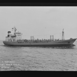 Schwarzweiß-Seitenansicht eines Frachtschiffs auf ruhiger See mit sichtbaren Aufbauten, Ladegestängen und Filmrand; Wasserzeichen des Internationalen Maritimen Museums Hamburg unten links.