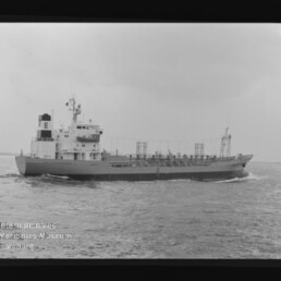 Tankschiff auf offenem Wasser in einer Schwarzweiß-Aufnahme: mittig positioniertes Schiff mit markantem Aufbauten und dem Buchstaben E auf dem Schornstein, sichtbaren Deckrohren und leichter Bugwelle, vor bedecktem Himmel; unten links Wasserzeichen des Internationalen Maritimen Museums Hamburg.