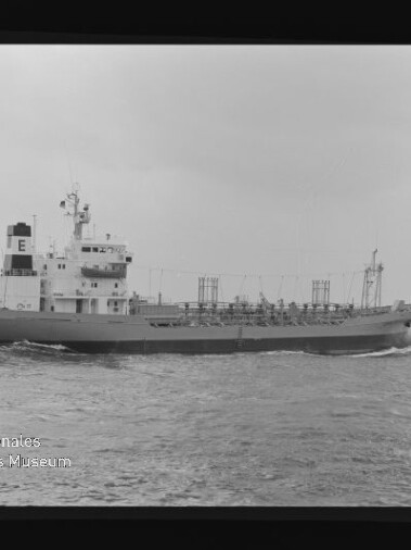 Tankschiff auf offenem Wasser in einer Schwarzweiß-Aufnahme: mittig positioniertes Schiff mit markantem Aufbauten und dem Buchstaben E auf dem Schornstein, sichtbaren Deckrohren und leichter Bugwelle, vor bedecktem Himmel; unten links Wasserzeichen des Internationalen Maritimen Museums Hamburg.