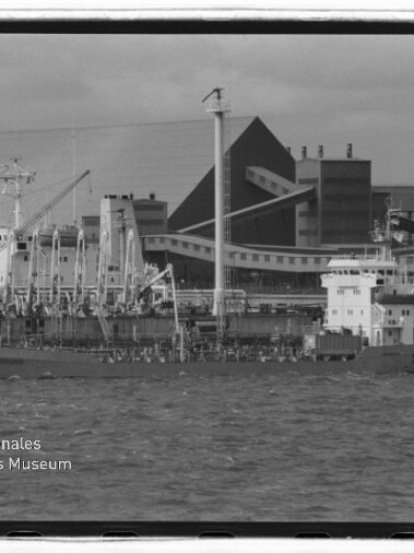Schwarz-weiß-Foto eines länglichen Frachtschiffs am Industriehafen mit zahlreichen Kränen und großen Lagergebäuden im Hintergrund, welliges Wasser im Vordergrund und Museumswasserzeichen unten links.