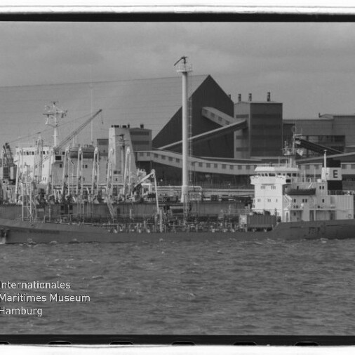 Schwarz-weiß-Foto eines länglichen Frachtschiffs am Industriehafen mit zahlreichen Kränen und großen Lagergebäuden im Hintergrund, welliges Wasser im Vordergrund und Museumswasserzeichen unten links.