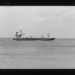 Schwarzweiß-Foto eines Frachtschiffs auf offener See, das von rechts nach links fährt; niedriger Aufbautenblock mittschiffs, ruhige Wellen und wolkiger Himmel, mit Wasserzeichen 