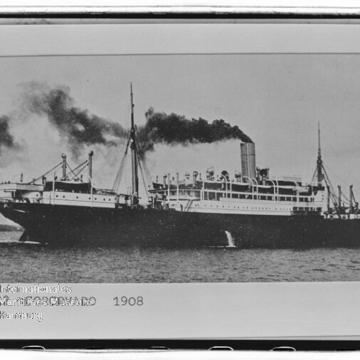 Schwarzweiß-Foto eines großen Dampfschiffs auf ruhiger See von 1908, mit mittigem Schornstein, dunklem Rauch, mehreren Masten und der Jahresangabe „1908“ unten im Bild.