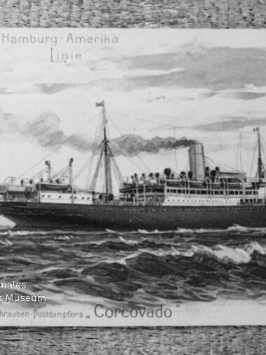 Schwarzweiß-Postkarte mit einer Illustration des Dampfschiffs „Corcovado“ der Hamburg‑Amerika Linie, das mit rauchendem Schornstein und Masten durch bewegte See fährt; die Karte wird an den oberen Ecken gehalten und trägt ein Museumswasserzeichen.