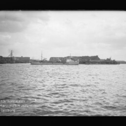 Schwarzweißaufnahme eines Hafens: wellige Wasserfläche im Vordergrund, mehrere Schiffe am Kai und lange Lagerhäuser an der Hafenpromenade unter einem bewölkten Himmel; Museumswasserzeichen unten links.