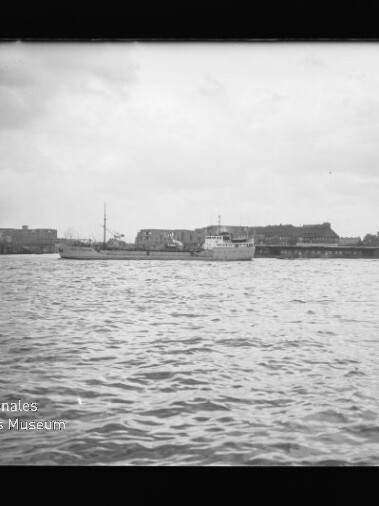 Schwarzweißaufnahme eines Hafens: wellige Wasserfläche im Vordergrund, mehrere Schiffe am Kai und lange Lagerhäuser an der Hafenpromenade unter einem bewölkten Himmel; Museumswasserzeichen unten links.