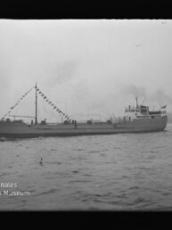 Schwarzweiß-Foto eines langen Frachtschiffs mit Girlanden aus Signalfahnen am Mast, rauchendem Schornstein und einem kleinen Schlepper neben dem Heck, im Vordergrund welliges Wasser mit einem einzelnen Vogel und im Hintergrund eine neblige Hafenkulisse; Museumswasserzeichen links unten.