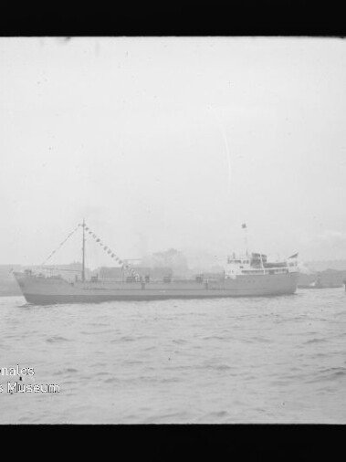 Schwarzweißaufnahme eines mittleren Frachtschiffs mit aufgehängten Wimpelketten, das auf ruhigem Wasser vor einer Uferlinie fährt; rechts ein kleiner Schlepper und unten links ein Wasserzeichen des Internationalen Maritimen Museums Hamburg.