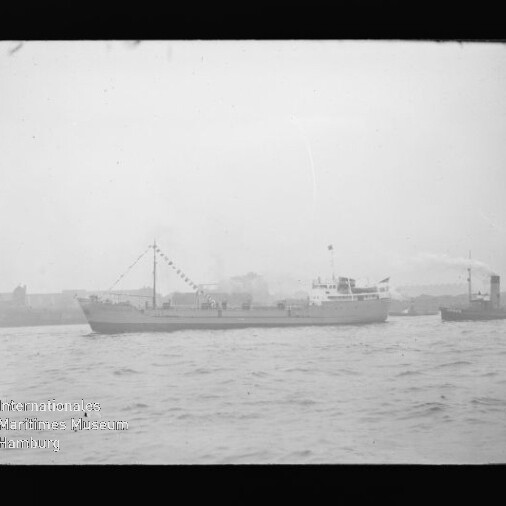 Schwarzweißaufnahme eines mittleren Frachtschiffs mit aufgehängten Wimpelketten, das auf ruhigem Wasser vor einer Uferlinie fährt; rechts ein kleiner Schlepper und unten links ein Wasserzeichen des Internationalen Maritimen Museums Hamburg.
