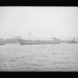 Schwarzweißaufnahme eines Frachtschiffs mit geschmücktem Mast und Wimpelketten, das in einem Industriehafen vor welligem Wasser vorbeifährt; im Hintergrund Hafenbauten und unten links der Schriftzug Internationales Maritimes Museum Hamburg.