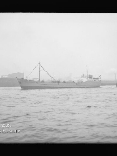 Schwarzweißaufnahme eines Frachtschiffs mit geschmücktem Mast und Wimpelketten, das in einem Industriehafen vor welligem Wasser vorbeifährt; im Hintergrund Hafenbauten und unten links der Schriftzug Internationales Maritimes Museum Hamburg.