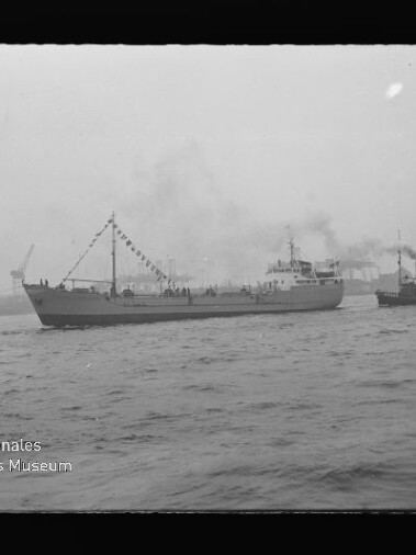 Schwarzweißaufnahme eines beflaggeten Frachtschiffs, das von einem Schlepper durch einen nebligen Industriehafen mit rauchenden Schornsteinen und Kränen begleitet wird; unten links das Logo des Internationalen Maritimen Museums Hamburg.