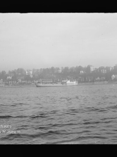 Schwarzweiß-Foto eines Frachtschiffs auf ruhiger See vor einer Uferlinie mit Häusern und Bäumen, unten links sichtbar das Wasserzeichen/Logo des Internationalen Maritimen Museums Hamburg.