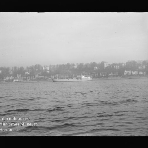 Schwarzweiß-Foto eines Frachtschiffs auf ruhiger See vor einer Uferlinie mit Häusern und Bäumen, unten links sichtbar das Wasserzeichen/Logo des Internationalen Maritimen Museums Hamburg.