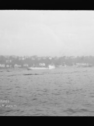 Schwarz-weiß-Aufnahme einer weiten Wasserfläche mit einem sich rechts der Bildmitte bewegenden Passagierboot und einigen Ruderbooten, im Hintergrund ein bewaldeter Uferstreifen mit Reihenhäusern und einem hellen, bewölkten Himmel; Museumslogo unten links.