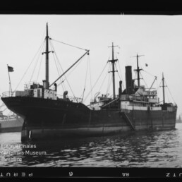 Schwarzweißfoto eines historischen Frachtdampfers mit Schornstein, mehreren Masten und Ladegeschirren, im Hafen vor ruhigem Wasser mit Spiegelung; unten links das Wasserzeichen 