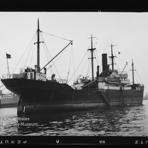 Schwarzweißfoto eines historischen Frachtdampfers mit Schornstein, mehreren Masten und Ladegeschirren, im Hafen vor ruhigem Wasser mit Spiegelung; unten links das Wasserzeichen 