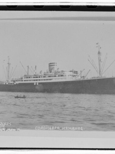 Schwarzweißfoto eines großen Passagier-/Frachtschiffs Cordillera (Hamburg) auf ruhigem Wasser, sichtbar sind Schornstein, Ladekräne und ein kleines Ruderboot im Vordergrund sowie ein Museumswasserzeichen in der linken unteren Ecke.