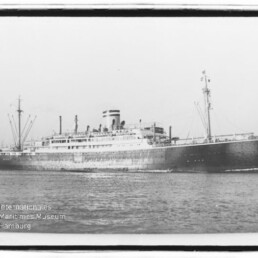 Schwarzweißfoto eines großen historischen Passagierschiffs mit einem einzelnen Schornstein und mehreren Lade- bzw. Masten, das auf ruhiger See entlangfährt; unten links sichtbares Museumssignet.