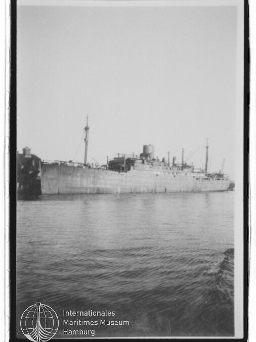 Schwarzweiß-Foto eines großen grauen Frachtschiffs mit Schornstein und mehreren Masten, längsseits an einem Kai bei ruhigem Wasser; unten links das Logo des Internationalen Maritimen Museums Hamburg.