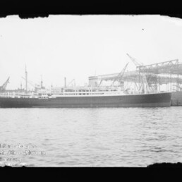 Schwarzweißfoto eines langgestreckten Passagierschiffs mit dunklem Rumpf und mehreren Aufbauten, vor einem Industrie-Kai mit großen Kränen und Wasser im Vordergrund.