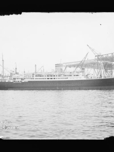 Schwarzweißfoto eines langgestreckten Passagierschiffs mit dunklem Rumpf und mehreren Aufbauten, vor einem Industrie-Kai mit großen Kränen und Wasser im Vordergrund.