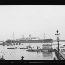 Schwarzweißaufnahme eines großen Passagierschiffs im Hafen, im Vordergrund zahlreiche kleine Boote, ein hölzerner Anleger mit einigen Personen, eine hohe Laterne am rechten Bildrand und ein Museumswasserzeichen in der linken unteren Ecke.