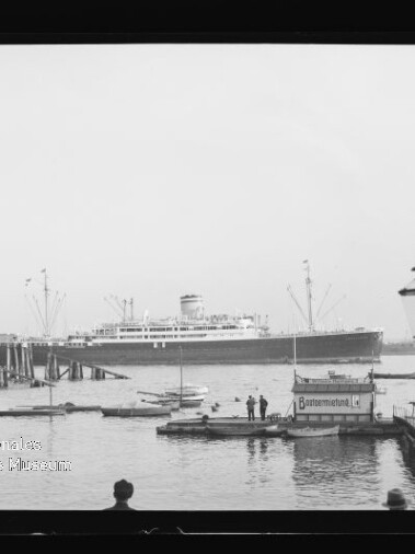 Schwarzweißaufnahme eines großen Passagierschiffs im Hafen, im Vordergrund zahlreiche kleine Boote, ein hölzerner Anleger mit einigen Personen, eine hohe Laterne am rechten Bildrand und ein Museumswasserzeichen in der linken unteren Ecke.