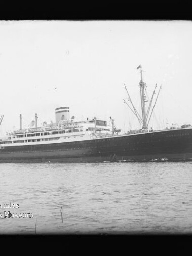 Schwarzweiß-Foto eines großen Passagierdampfers im Seitenprofil auf ruhiger See vor einem Hafen, mit hohem Schornstein, mehreren Ladebäumen und einer Flagge am Heck; unten links ein runder Museumsstempel und ein schwarzer Negativrahmen um das Bild.
