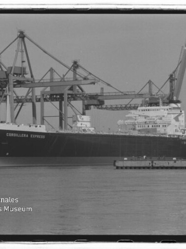 Großes Frachtschiff „Cordillera Express“ liegt am Kai eines Containerterminals vor mehreren hohen Verladekränen, mit ruhiger Wasserfläche im Vordergrund und einem Museumslogo unten links.