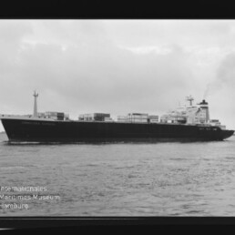 Schwarz-weiß-Foto eines großen Containerschiffs in Seitenansicht auf ruhiger See mit gestapelten Containerkästen, Rauch aus dem Schornstein, bewölktem Himmel sowie sichtbarem Filmrahmen und Museumswasserzeichen.