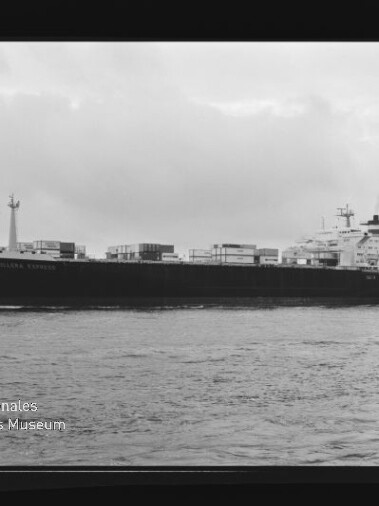 Schwarz-weiß-Foto eines großen Containerschiffs in Seitenansicht auf ruhiger See mit gestapelten Containerkästen, Rauch aus dem Schornstein, bewölktem Himmel sowie sichtbarem Filmrahmen und Museumswasserzeichen.
